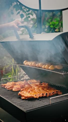 Juicy pork ribs grilling on lower rack, small potatoes roasting above inside smoky outdoor barbecue smoker.