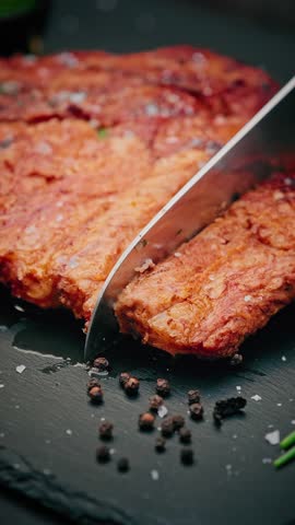Close-up of juicy grilled pork ribs being sliced with a knife, sprinkled with coarse salt and herbs, served with cherry tomato on black slate.