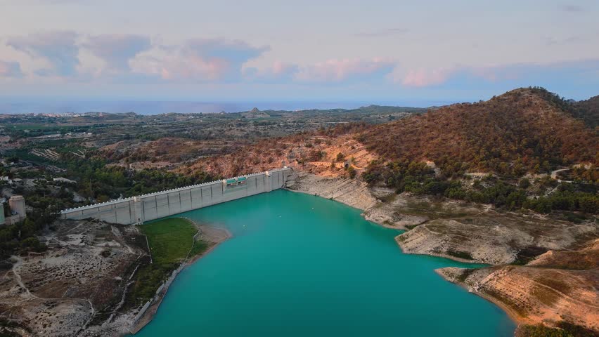 Drone video of a turquoise reservoir in Spain, showing the dam structure surrounded by hills and countryside under a soft pastel evening sky.