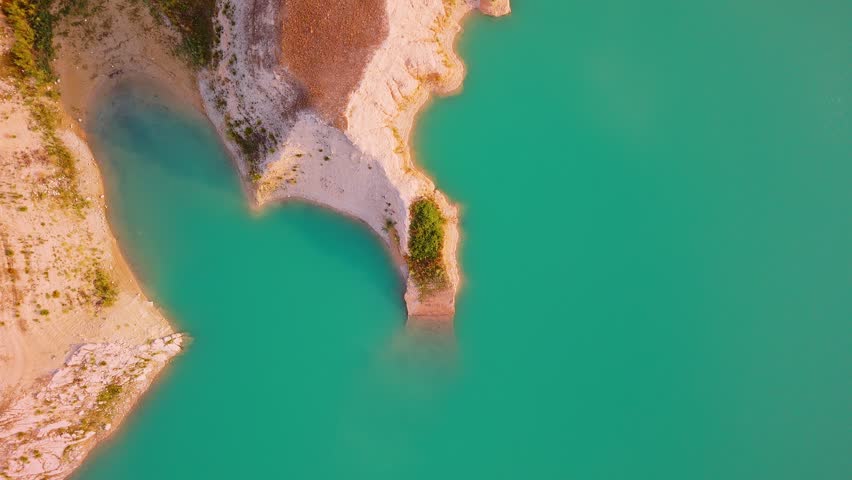 Drone video captures a rocky shoreline with green vegetation surrounded by bright turquoise waters of a lake in Spain, viewed directly from above.
