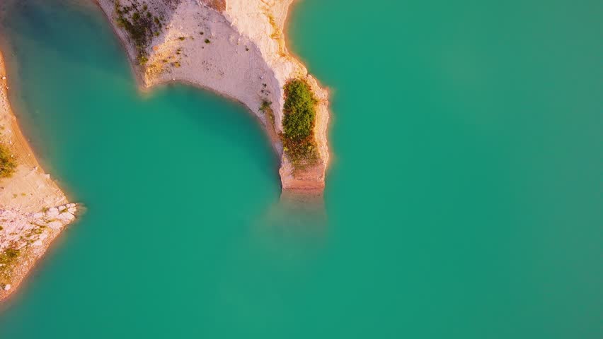 Drone video captures a rocky shoreline with green vegetation surrounded by bright turquoise waters of a lake in Spain, viewed directly from above.