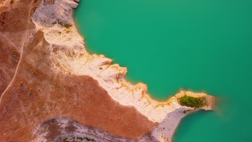 Drone video captures a rocky shoreline with green vegetation surrounded by bright turquoise waters of a lake in Spain, viewed directly from above.