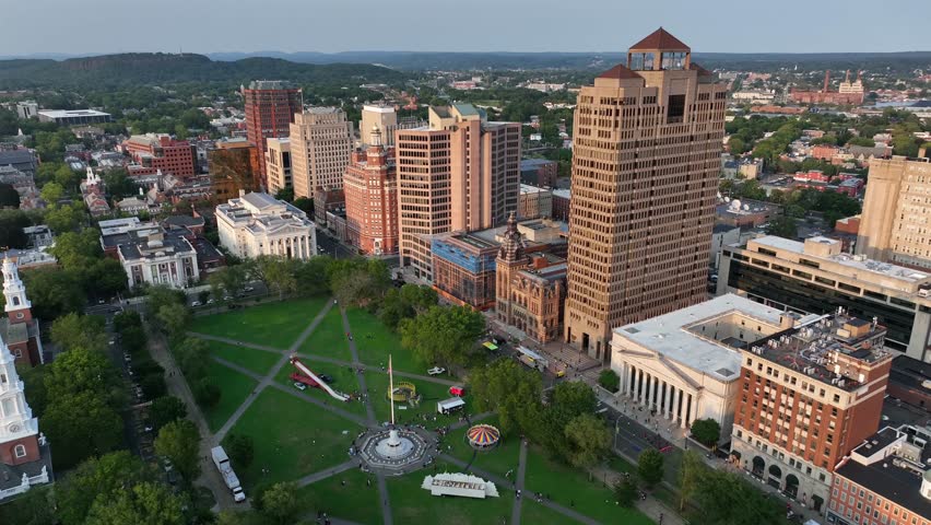 Patriotic American flag on flagpole. Downtown towers of New Haven town at golden sunset. Aerial top down shot. Green park area and skyscrapers in city. Connecticut, America.