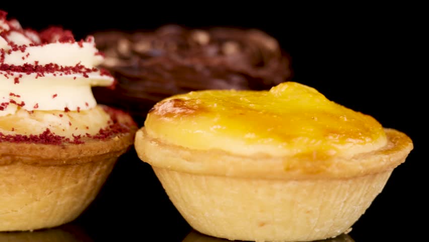 Close-up rotating view of assorted mini baked desserts, including chocolate cream tarts, egg tarts, and red velvet cupcakes, on a reflective black surface with soft studio lighting