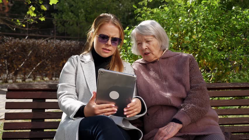 Portrait of a young adult granddaughter wearing a coat and sunglasses, holding a tablet, with her old grandmother sitting on a bench. They are both looking at the screen and talking. - Powered by Shutterstock - Get 15% off with code: PIKWIZARD15