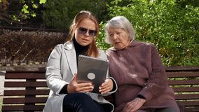 Portrait of a young adult granddaughter wearing a coat and sunglasses, holding a tablet, with her old grandmother sitting on a bench. They are both looking at the screen and talking. - Powered by Shutterstock - Get 15% off with code: PIKWIZARD15