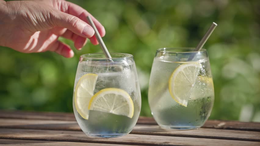 human hand put straw into glass of fresh sparkling water with ice and lemon slice on sunny green garden background, food closeup outdoor