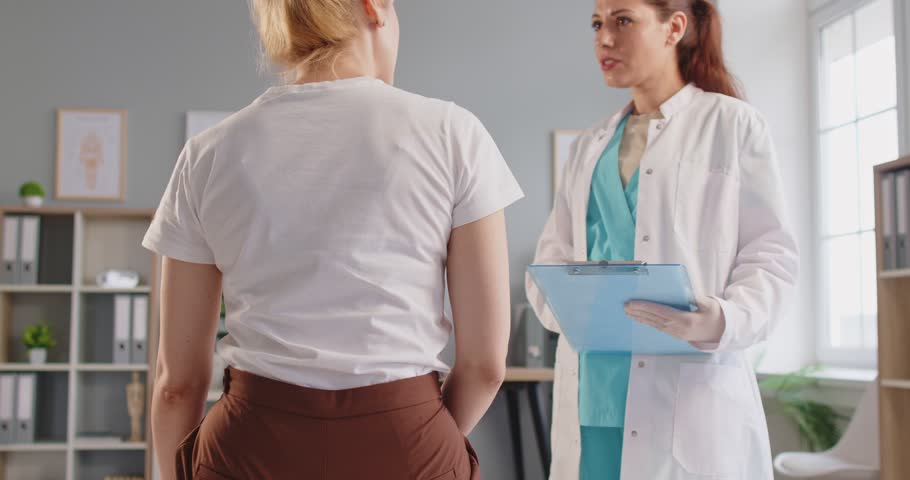 Young sick woman sitting on the couch in the doctor's office suffering from backache during medical examination in clinic. Physician female doctor listening to the patient's complaints. 4k video.
