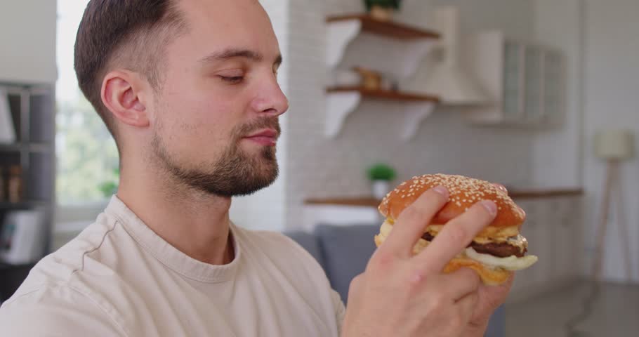 Man eating tasty burger, biting with big appetite, enjoying takeaway fast food, keeping eyes closed from pleasure, feeling hungry, posing indoor. Unhealthy eating and junk food concept