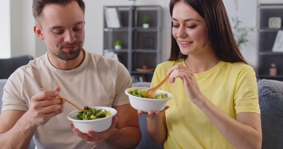 Close-up of young 30s man and woman holding white bowls, smiling and eating natural, nutritious, green, vitamin-rich salad seated on sofa at home. Keep healthy, vegetarian lifestyle concept