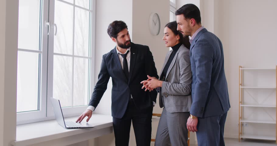Portrait of confident man professional realtor standing with couple clients in modern new apartment looking at laptop discussing contract buying property. Move, rent and real estate market concept.