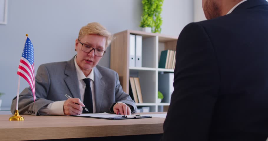 Male person sitting in the office of the US public services or embassy having consular visa interview with mature woman officer with US flag at the desk of official's workplace. 4k video.