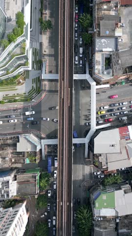 Aerial top-down view of BTS Skytrain on elevated railway tracks above busy street in Bangkok, Thailand. Urban transportation scene with modern train, traffic, and city buildings.