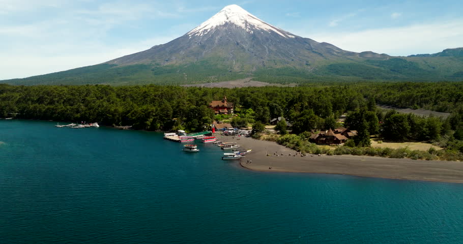 Forward-moving drone glides across Lake Todos los Santos toward a small harbor, as snowcapped Osorno volcano rises beyond forest and dark sand shore beneath a clear summer sky.