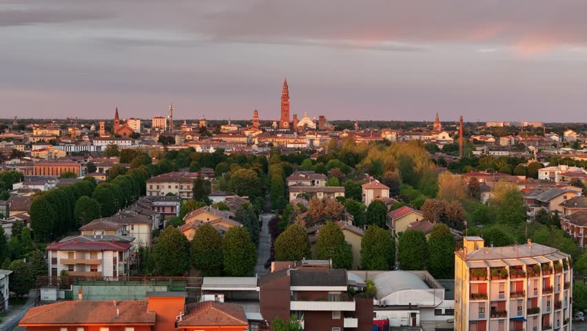 Drone ascends above Cremona, revealing tree-lined avenues, historic rooftops, and iconic landmarks including the Cathedral of Cremona and the towering Torrazzo, bathed in golden sunset light.