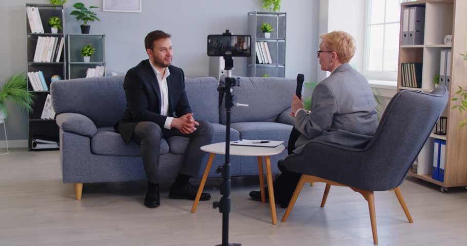 Young male company CEO speaker sitting on sofa in office workplace setting discusses business, work, finance questions with female media program host, phone on tripod recording digital interview video