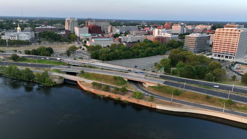 Aerial wide shot showing driving cars on interstate road in Trenton along Delaware River at sunset. Downtown with towers and blocks in background. New Jersey, USA in summer.