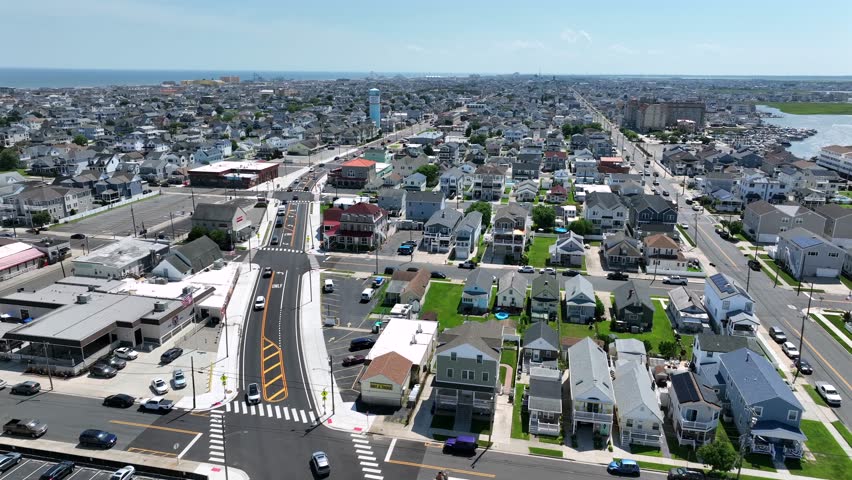 Aerial rising wide shot showing Wildwood City in New Jersey with Atlantic ocean in background. Sunny summer day with cars on main street in neighborhood. Beach houses and apartment blocks.