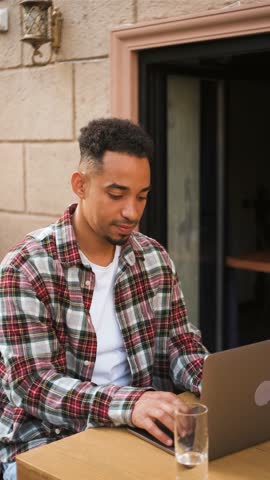 Young man working on a laptop outdoors at a modern cafe in the afternoon sunshine, focused on a creative project while enjoying a refreshing drink,vertical video