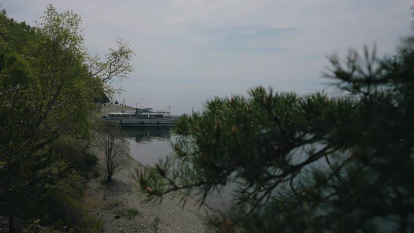 Passenger ferry approaching wooden pier, tourists anticipating arrival against verdant landscape at scenic lake Baikal shoreline. Concept of vacation and travel in Russia. Shooting in slow motion.