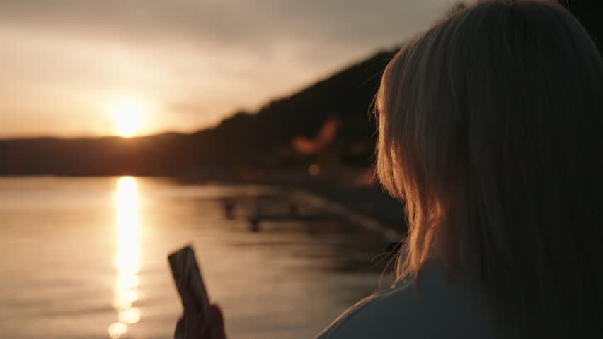 Back view of blonde woman talking on mobile phone standing near shore, enjoying peaceful atmosphere of setting sun over calm sea during golden hour, close-up. Tracking shot in slow motion.