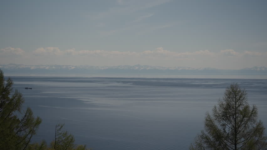 Cargo ship sailing on tranquil lake Baikal with snowy Sayan mountains in background, creating serene and picturesque scene under cloudy sky with pine trees, no people, nobody. Shooting in slow motion.