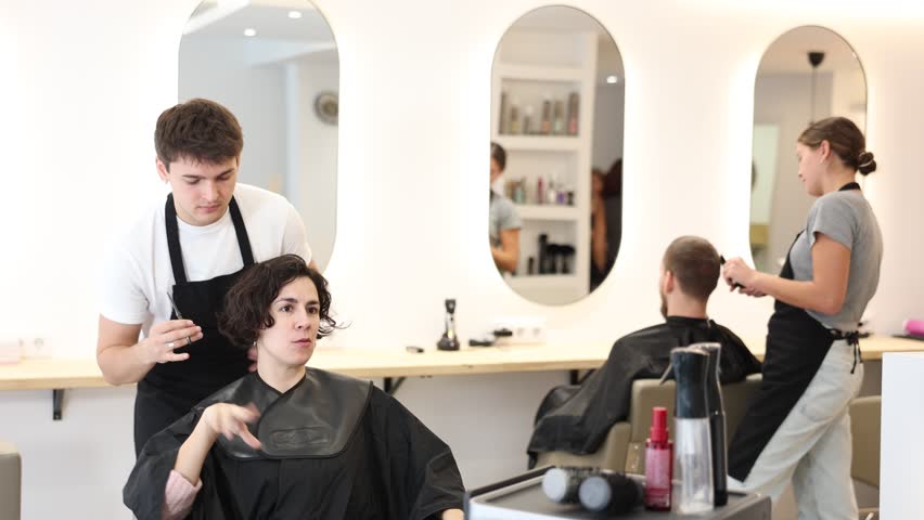Young guy hairdresser stylist gives haircut to adult woman client in beauty salon