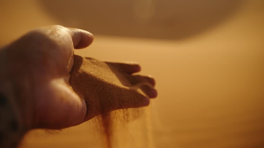 Close-up of golden desert sand slipping gracefully through a hand at sunset in the Sahara Desert, Morocco, with warm light creating a soft glow.