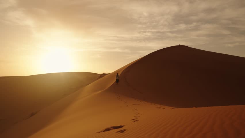 A traveler walks along the ridge of a golden sand dune in the Sahara Desert at sunrise, leaving footprints under the soft, warm glow of the Moroccan morning.