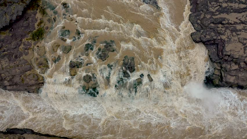 A top-down drone view of muddy Yellow River water crashing through rocks at Hukou Waterfall in China narrow gorge