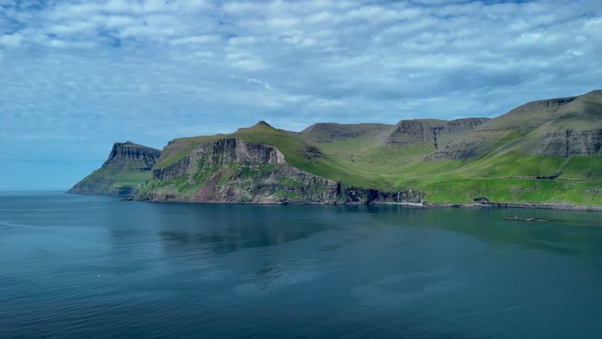 Stunning aerial view showcases the dramatic coastal cliffs and calm waters of the Faroe Islands. The vibrant green landscape harmonizes with the blue sea under a clear sky.