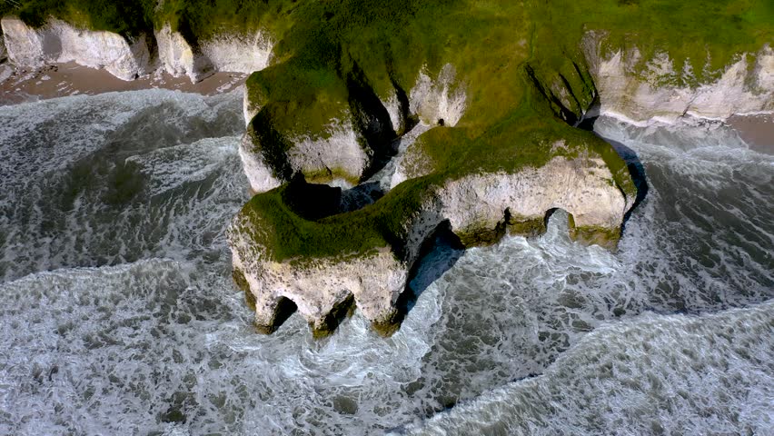 A drone view of steep white chalk cliffs and green-topped rocks meeting the waves at Flamborough Head, England
