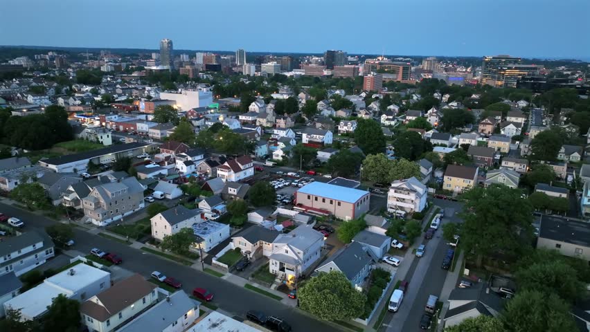 Peaceful suburb neighborhood with houses and homes at dusk. American town in summer with green trees. Downtown buildings and towers in background. Aerial flyover shot. Stamford, Connecticut. Wide shot