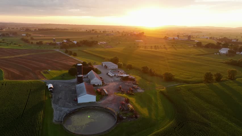 Large American farmstead with silo storage, barn and fields during bright golden sunset. Circular pond in rural suburb area of town. Aerial wide shot. Midwest, USA. Peaceful countryside scene.
