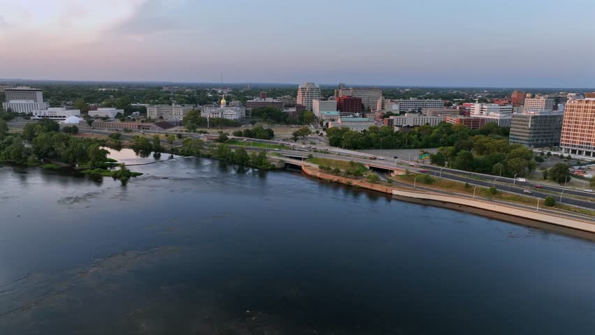 Aerial flyover river in Trenton city at sunset time. Tranquil waters and traffic on shoreline road. Downtown with towers and buildings in background. Wide shot. New Jersey in summer season.