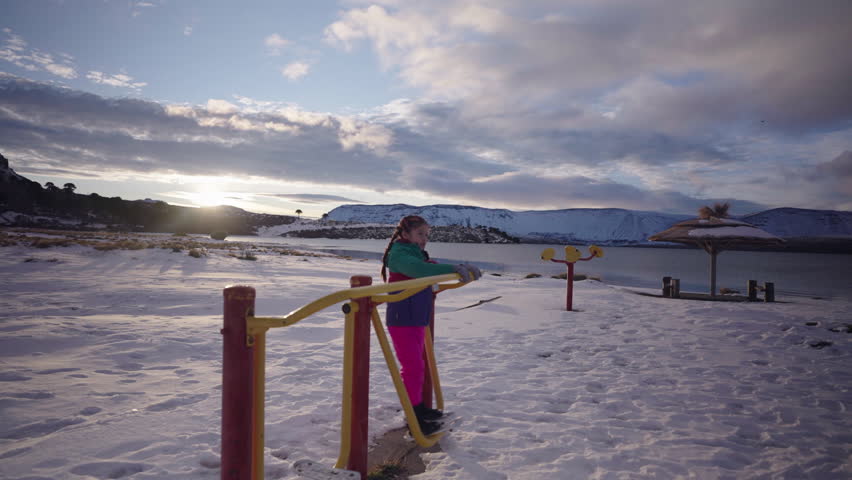 Young girl in colorful winter clothing plays on an outdoor ski simulator exercise machine beside a frozen lakeshore with mountains in Patagonia, Argentina, captured in wide ground-level shot.
