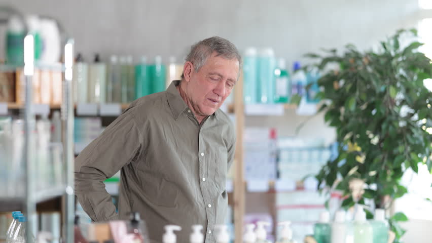 Elderly man is standing in a pharmacy and holding on to his back. Buyer came for an ointment for the lower back and joints. High quality 4k footage