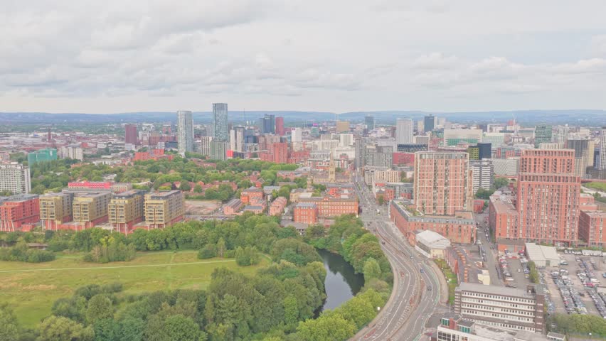 Drone pan over Salford highlights the River Irwell curving through green spaces, Trinity Way roadway, and Manchester’s skyline with Beetham Tower, Deansgate Square, and modern high-rises.