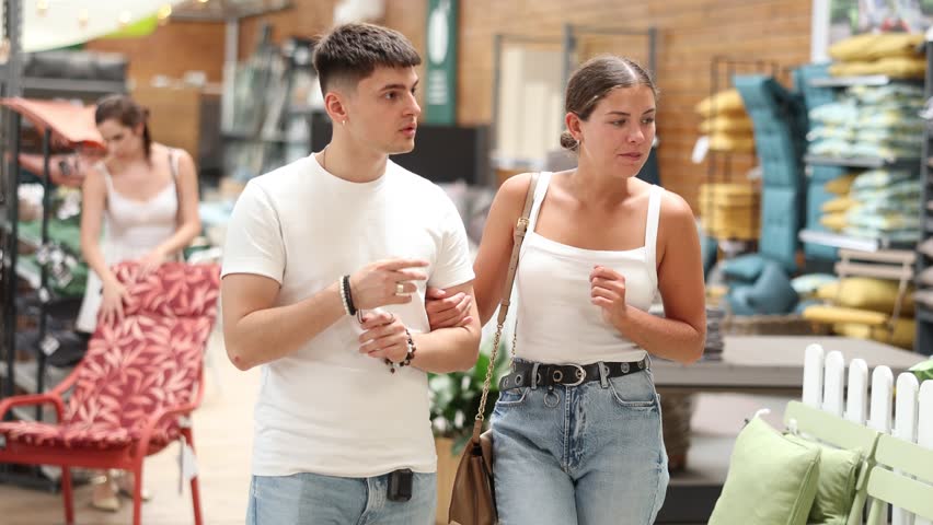 Carefree cheerful young couple engaging in shopping, walking down aisle of home and garden store, exploring with interest furniture and accessories around