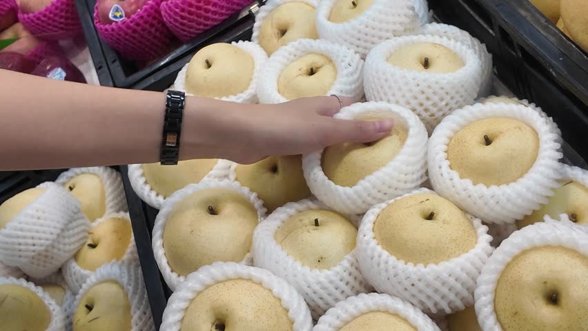 A person is holding a white plastic wrapped apple in a store. The pear are in a bin and are surrounded by plastic