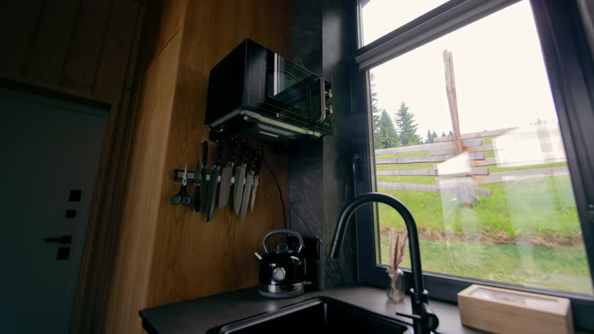 Modern cabin kitchen with forest view window. Compact and stylish cabin kitchen with black sink, microwave, and knife rack, offering a scenic view of trees and grassy outdoors.