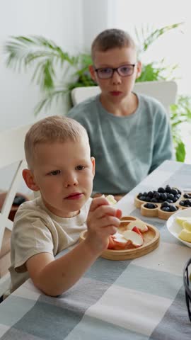 Two young brothers eating healthy breakfast with fresh fruits berries and apple slices at home kitchen table with plants. Concept of kids nutrition, family meals and healthy eating habits for children