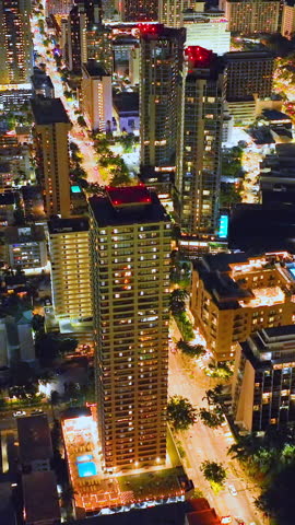 Glowing night streets of Honolulu on the island of Oahu. Tall hotel buildings with lights in Waikiki. Cars on the road. Aerial view. Vertical video.