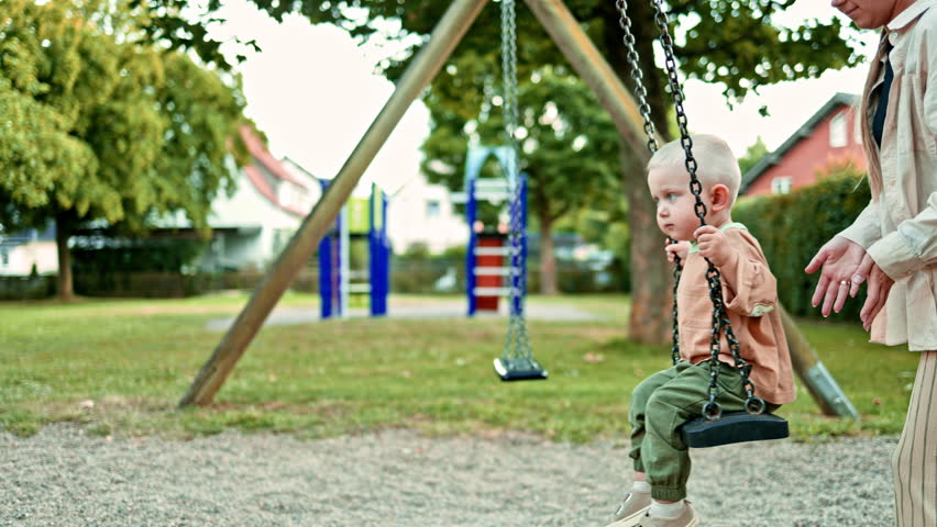 Mom on the playground swings her little son on a swing. Toddler sits on a swing and caring mother swings him. Mom and son walk in the fresh air. Happy family concept. Child development.