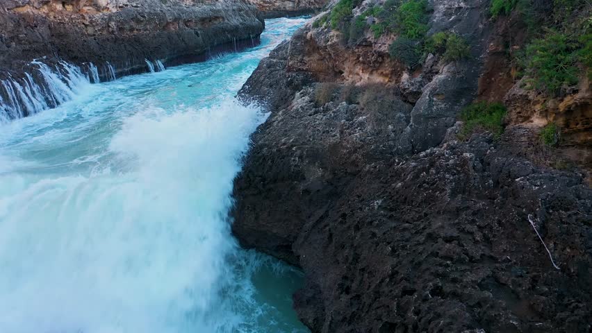 Powerful drone shot of a turbulent sea surging through a narrow rock channel. Waves crash and create small waterfalls, showcasing the raw, untamed force of nature in a dramatic close-up.