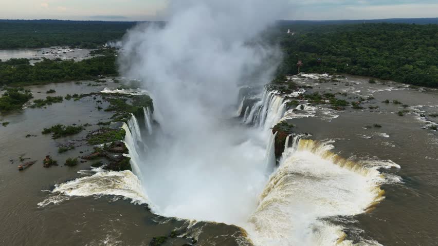 Massive volume of water crashes into Devil's Throat abyss Plume of vapor surges upward Drone motion reveals raw natural intensity as liquid thunder spirals into the vaporous void. Slow motion