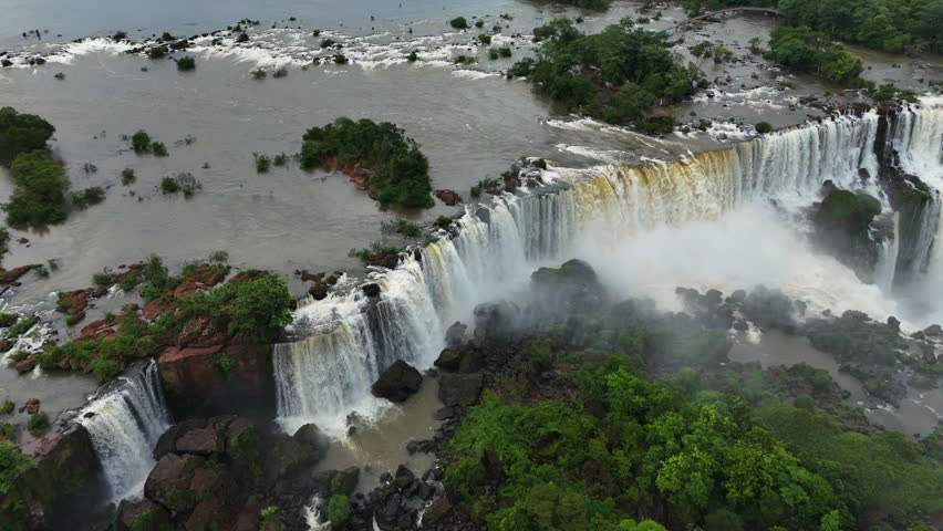 Powerful streams of Iguazu Falls rush over steep cliffs in Brazil creating heavy mist. Aerial movement captures the immense natural force. Water flows between islands and green forests below
