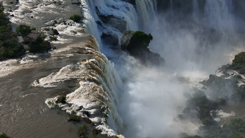 Aerial view of Iguazu Falls shows muddy water rushing over cliffs. The wild river carves through rainforest White spray rises from the deep below. The drone tracks the untamed force of nature