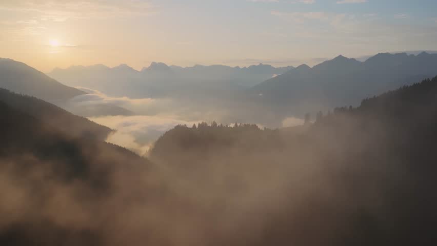 Breathtaking view of a golden sunrise over a vast mountain range. A sea of fog fills the valleys below, with a small village visible on a distant hill, creating a serene and majestic landscape.