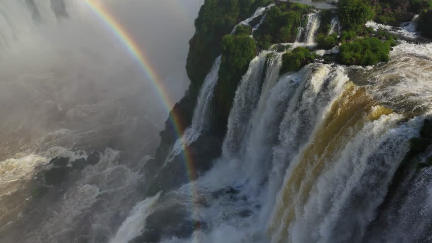 Aerial view slow motion video shows rainbow forming in thick mist at Iguazu Falls, Brazil Water plunges from green cliffs, meeting swirling clouds below Vivid colors create a magical natural scene